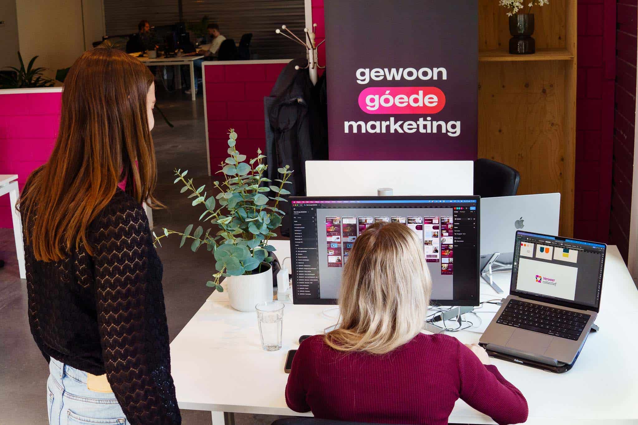 BURO210 Fullservice Marketingbureau Twee vrouwen werken samen op kantoor aan een home website ontwerp, met computers en marketing materialen op de tafel. Een creatieve en productieve werkomgeving.